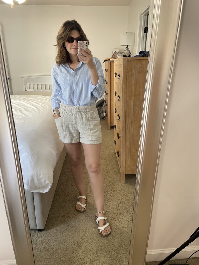 Outfit photo of blue and white strip button down, beige linen shorts, brown sunglasses, and white sandals