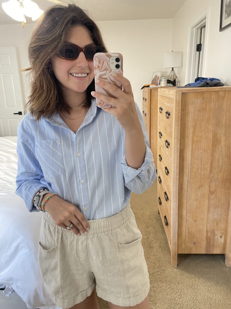 Upclose mirror picture of outfit, blue and white strip button down top, and beige linen shorts, with brown rectangle sunglasses 
