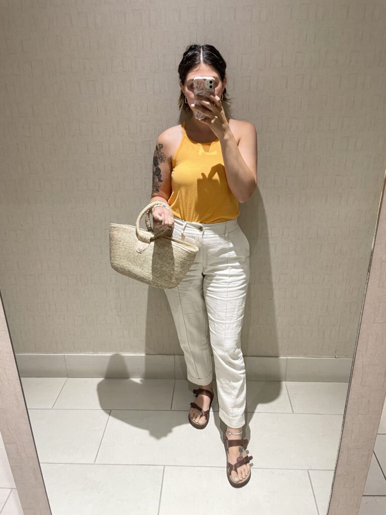 Outfit picture in a mirror, orange tank top with cream colored pants, brown sandals and straw bag 